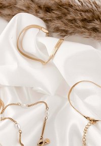 Aranylánc karkötő lapos szemekkel és gyöngygyöngyös láncnyaklánc, fehér szatén anyagon elrendezve, háttérben szárított pampafűvel.