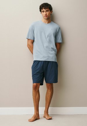 Junger Mann steht barfuß an der Wand, trägt ein hellblaues T-Shirt und marineblaue Shorts, die Hände hinter dem Rücken, neutraler Gesichtsausdruck.