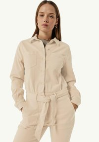 Chemise beige légère à manches longues avec des boutons, deux poches poitrine et une taille ceinturée. Tissu texturé et coupe décontractée.
