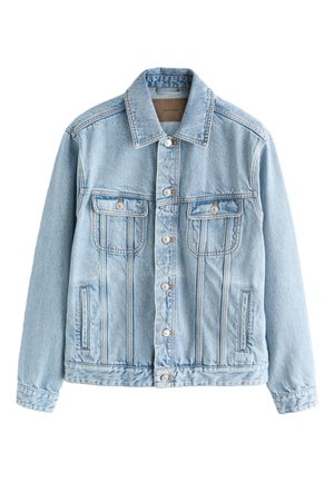 Junger Mann, der draußen steht und eine hellblaue Jeansjacke, ein gestreiftes Hemd, graue Hose und schwarze Schuhe neben einer gefliesten Wand und einem Metallpfosten trägt.