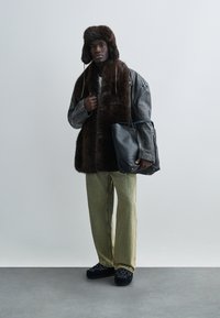 Brown fur-lined hat, gray leather jacket over a fur vest, green striped pants, and black shoes with floral patterns, holding a black bag.