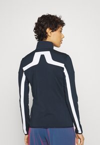 Giacca a maniche lunghe blu navy con strisce bianche a contrasto, colletto alto, design aderente e texture liscia. Versatile per abbigliamento sportivo.