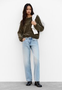 Maglione texturizzato verde oliva, jeans a gamba dritta blu chiaro e stivaletti neri con tacco. Top a righe bianche appoggiato sul braccio.