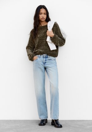 Maglione texturizzato verde oliva, jeans a gamba dritta blu chiaro e stivaletti neri con tacco. Top a righe bianche appoggiato sul braccio.