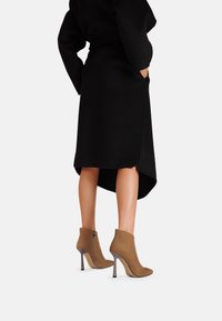 Stivaletti in suede marrone con tacco a stiletto, abbinati a una gonna nera con orlo asimmetrico e tasche laterali.