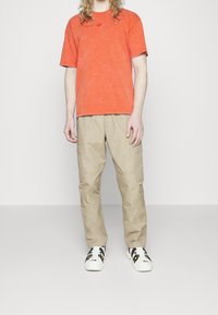 Orange bomullströja med rund hals och en liten logotyp. Beige cargobyxor med fickor. Svart-och-vit sneakers med ränder.