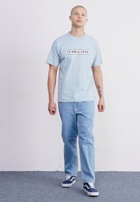 T-shirt en coton bleu clair avec un logo "VOLCOM" noir, associé à un jean en denim bleu clair. Les baskets présentent une combinaison de couleurs marine et blanche.