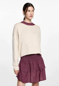 Beige verkürzter Pullover, kombiniert mit einem weinroten, blumengemusterten Smock-Rock. Der Pullover hat einen runden Halsausschnitt, während der Rock rüschierte Lagen aufweist.