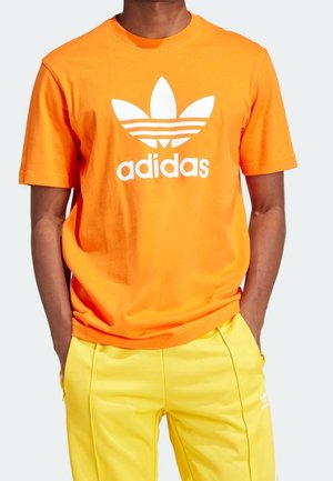 Orange Baumwoll-T-Shirt mit großem weißem Adidas-Logo und Grafik mit drei Streifen. Kombiniert mit gelben Jogginghosen aus glattem Material.