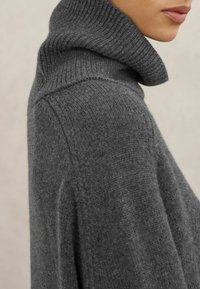 Grijze gebreide trui met een hoge, geribbelde kraag, voorzien van een zachte textuur en aflopende schouders, die een ontspannen pasvorm en strakke lijnen benadrukt.