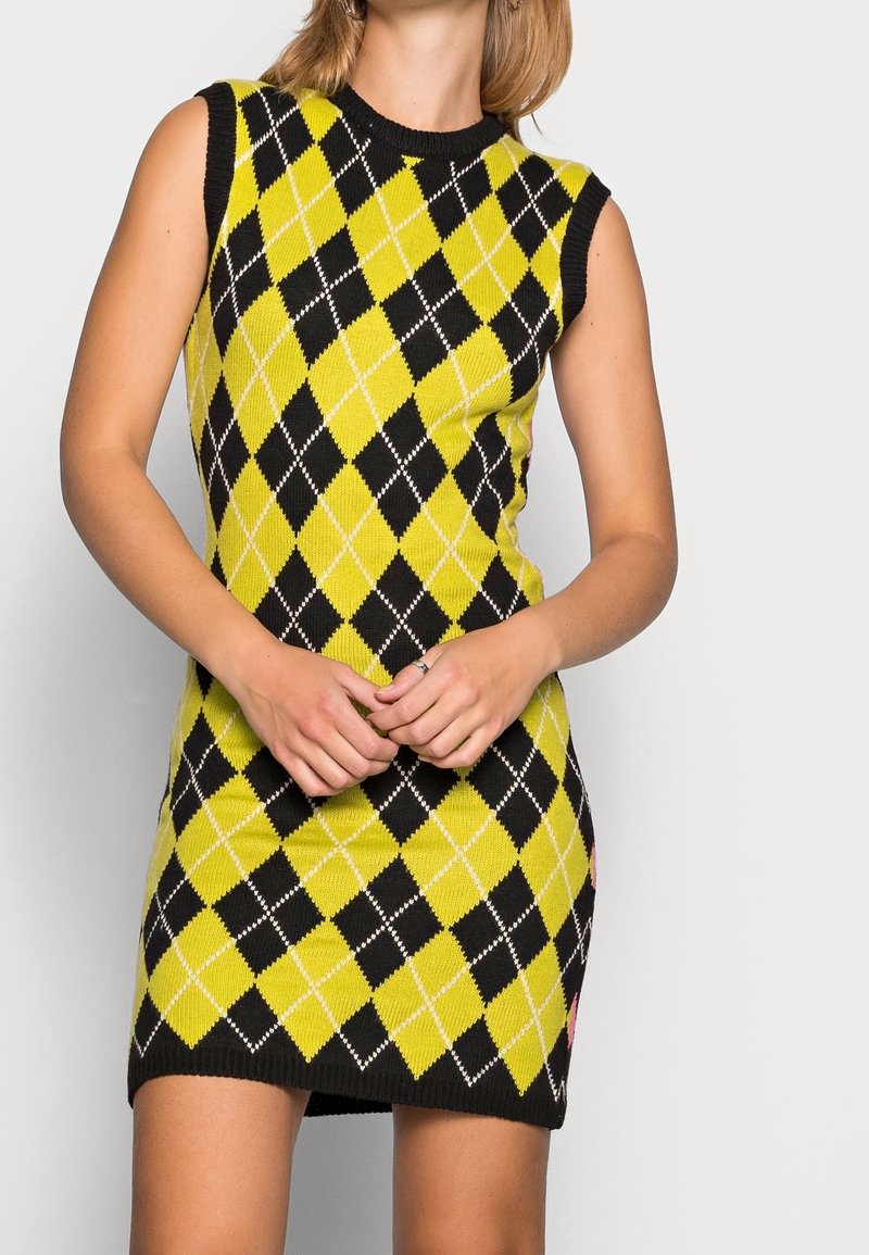 Femme portant une robe tricotée sans manches jaune et noire à motif losange, mains croisées à la taille.
