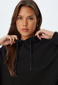 Sudadera negra con capucha, que cuenta con cordón, textura de tela suave y puños acanalados. El modelo está de frente con el cabello cayendo sobre los hombros.