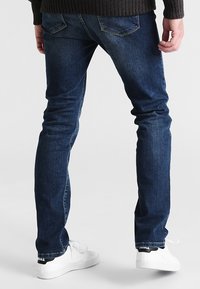 Jeans de denim azul oscuro con corte slim, que presentan un sutil desteñido, dos bolsillos traseros y bordes desgastados, combinados con zapatillas blancas.