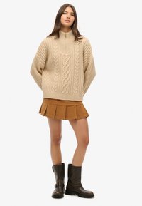 Beige chunky strikket sweater med kabelmønster; sammen med en kort brun plisseret nederdel og knæhøje sorte læderstøvler.