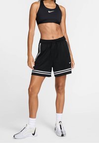 Sostén deportivo negro de Nike con diseño de espalda cruzada, combinado con pantalones cortos negros con rayas blancas. Completado con zapatillas blancas y calcetas hasta el tobillo.