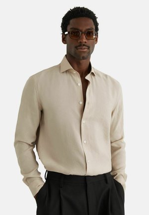 Mees, kes kannab beeži nööpidega särki, musti pükse ja toonitud ristkülikukujulisi prille, käed taskutes, valgel taustal.