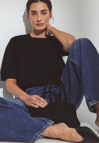 Femme assise contre un mur blanc, portant un haut noir, un jean bleu large, des ballerines noires pointues, tenant un sac à main noir sur le sol.