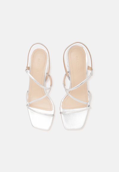 Paire de sandales argentées à brides ouvertes avec bout carré, fines lanières croisées et semelles intérieures beiges sur un fond clair uni.
