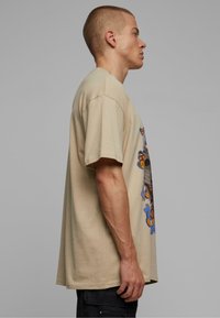 Camiseta de algodón beige con mangas cortas, que presenta un diseño gráfico frontal de mariposas y flores en azul y naranja. Vista lateral.