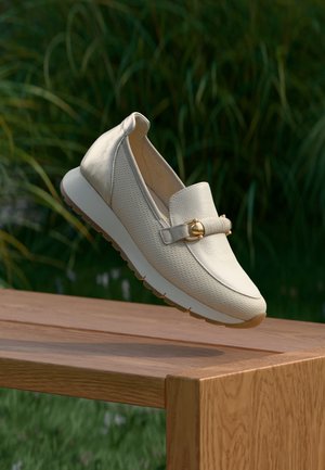 Béžové kožené mokasíny s textúrovaným povrchom a zlatým akcentom ležia na drevenej lavičke, majú hrubú bielu podrážku a zdvihnutý zadok.