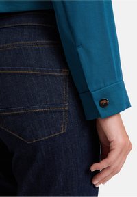 Teal long-sleeve shirt with buttoned cuff, paired with dark denim jeans featuring orange stitching, highlighting pocket detail and texture.