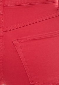 Tela de mezclilla roja con un primer plano de un bolsillo trasero y detalles de costura. El material tiene una textura suave con líneas de costura bien definidas.