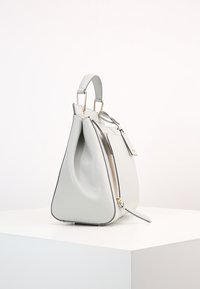 Biała skórzana torebka z górnym uchwytem i bocznym zamkiem błyskawicznym, wyeksponowana na białym postumencie na jednolitym tle.