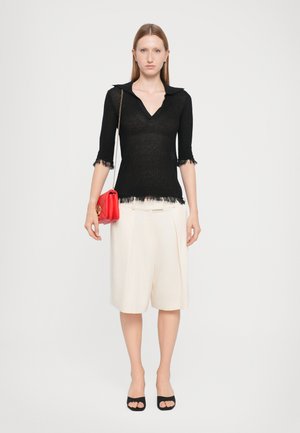 Schwarzes Stricktop mit Kragen und ausgefransten Ärmeln, kombiniert mit knielangen cremefarbenen Shorts und schwarzen offenen High Heels. Rote Handtasche mit Kettenriemen.