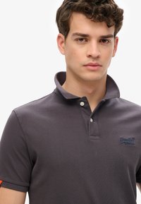 Grå polo t-shirt lavet af tekstureret stof, med kravehals, tre knapper og et broderet logo på brystet.