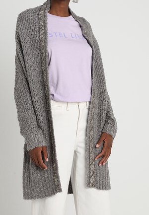 Personne portant un cardigan en tricot gris texturé sur un t-shirt imprimé violet pastel et un pantalon blanc taille haute sur un fond uni.