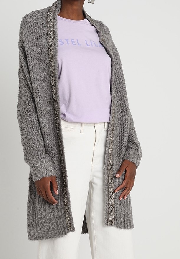 Personne portant un cardigan en tricot gris texturé sur un t-shirt imprimé violet pastel et un pantalon blanc taille haute sur un fond uni.