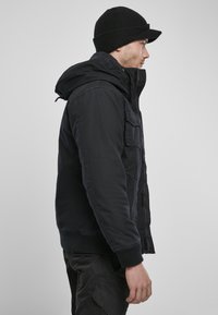 Černá kapucová bunda se zipem vpředu, našitými kapsami a žebrovanými manžetami. Nosí se s odpovídající černou zimní čepicí. Jednoduchý design, funkční.