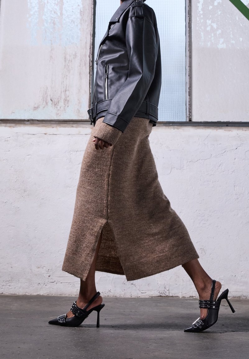 Personne marchant à l'intérieur portant une veste en cuir noire, une longue jupe marron tricotée avec une fente, et des chaussures slingback à talons hauts noires.