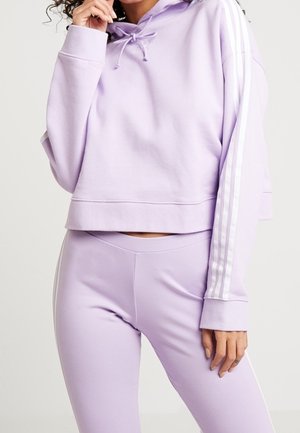 Vrouw draagt een bijpassende lila hoodie en leggings met witte strepen op de mouwen, staand met één hand dicht bij haar gezicht.