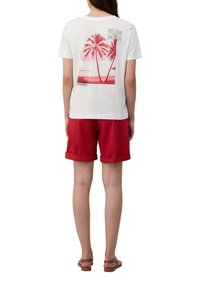 T-shirt blanche avec un graphique tropical rose comprenant des palmiers et un texte au dos, assortie à un short rouge et des sandales marron.