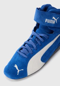 Plave swede visoke tenisice s zakrivljenim dizajnom bijele boje sa strane i plavim vezicama. Imaju podstavljeni ovratnik oko gležnja i Puma logo na remenu.