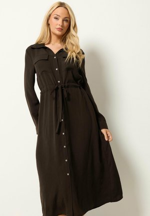 Frau trägt ein langärmeliges schwarzes Kleid mit Knöpfen und Taillengürtel, steht vor einem schlichten hellen Hintergrund.