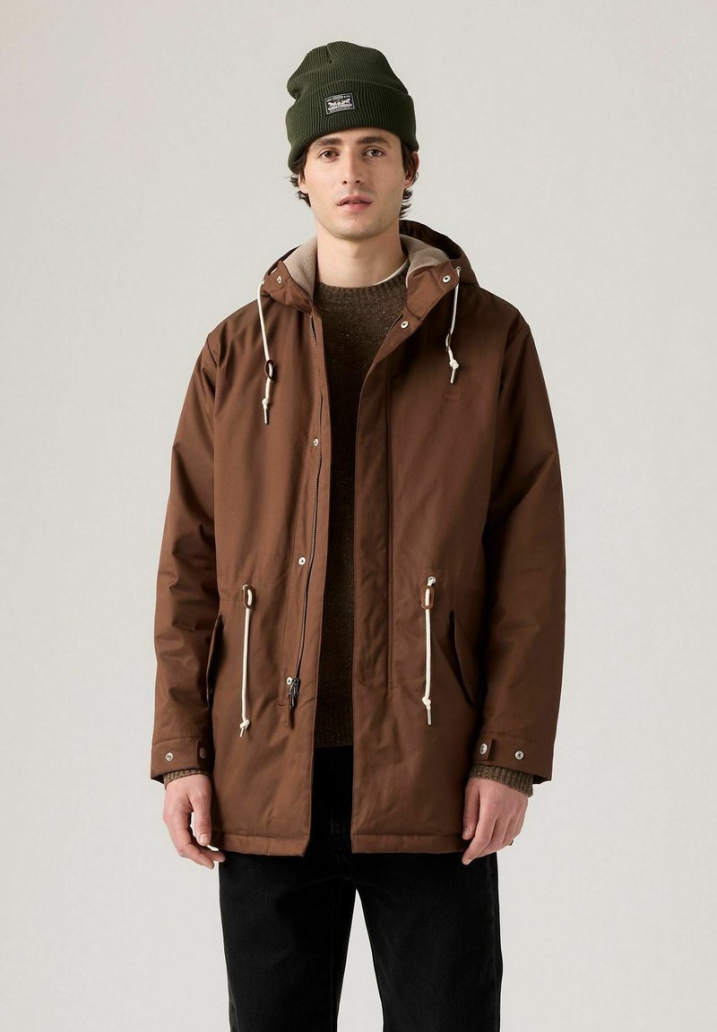 Brun vattentät jacka med huva, dragsko-stängningar och två framfickor. Bärs över en brun tröja och matchas med en grön mössa.