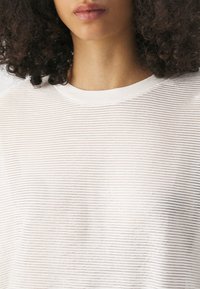 Gros plan d'une personne aux cheveux bouclés portant un t-shirt rayé horizontalement blanc et gris clair avec un col rond.