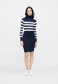 Μπλε σκούρο και λευκό ριγέ πλεκτό φόρεμα με ψηλό γιακά, εφαρμοστή σιλουέτα, μακριά μανίκια και μήκος έως το γόνατο. Φορεμένο με λευκά αθλητικά παπούτσια.