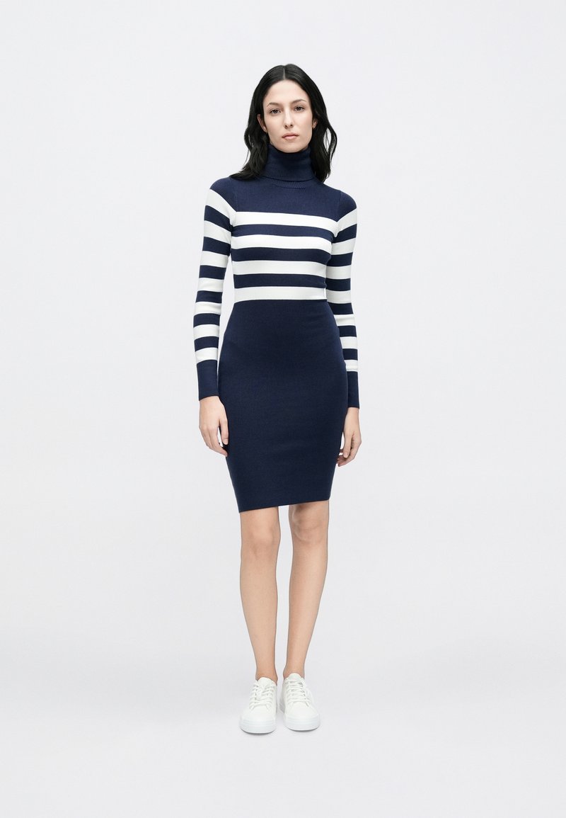 Marineblau-weiß gestreiftes Strickkleid mit Rollkragen, figurbetonter Silhouette, langen Ärmeln und knielangem Saum. Kombiniert mit weißen Sneakers.