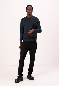 Dunkelblauer Sweatshirt, schwarze Nadelstreifen-Hose und schwarze klobige Stiefel. Eine kleine schwarze Umhängetasche wird über die Schulter getragen.