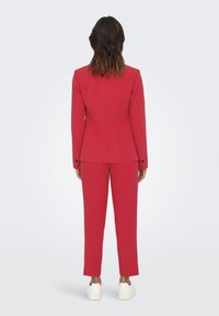 Costume rouge avec blazer ajusté et pantalon assorti, présentant une texture lisse et un design minimaliste, associé à des baskets blanches.