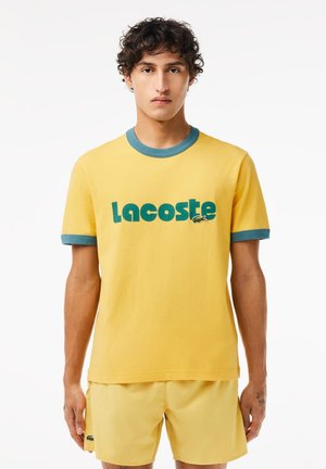 Camiseta de algodón amarilla con cuello y puños acanalados en azul. Presenta un audaz logo verde de "Lacoste" y un gráfico de cocodrilo. Diseño simple y casual.