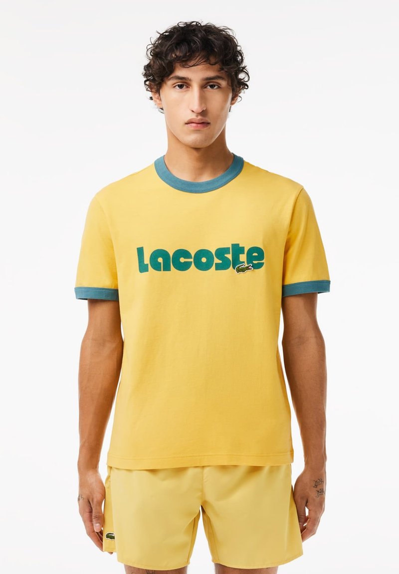 Żółta bawełniana koszulka z niebieską żebrowaną kołnierzykiem i mankietami. Posiada wyraziste zielone logo "Lacoste" oraz grafikę krokodyla. Prosty, casualowy design.