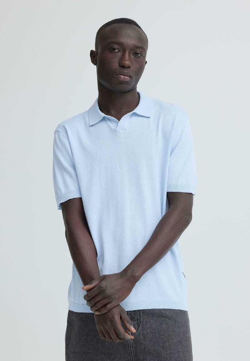 Άνδρας που φοράει γαλάζιο κοντομάνικο πόλο πουκάμισο και σκούρα γκρι παντελόνια, όρθιος με τα χέρια χαλαρά σταυρωμένα μπροστά σε απλό ανοιχτό φόντο.