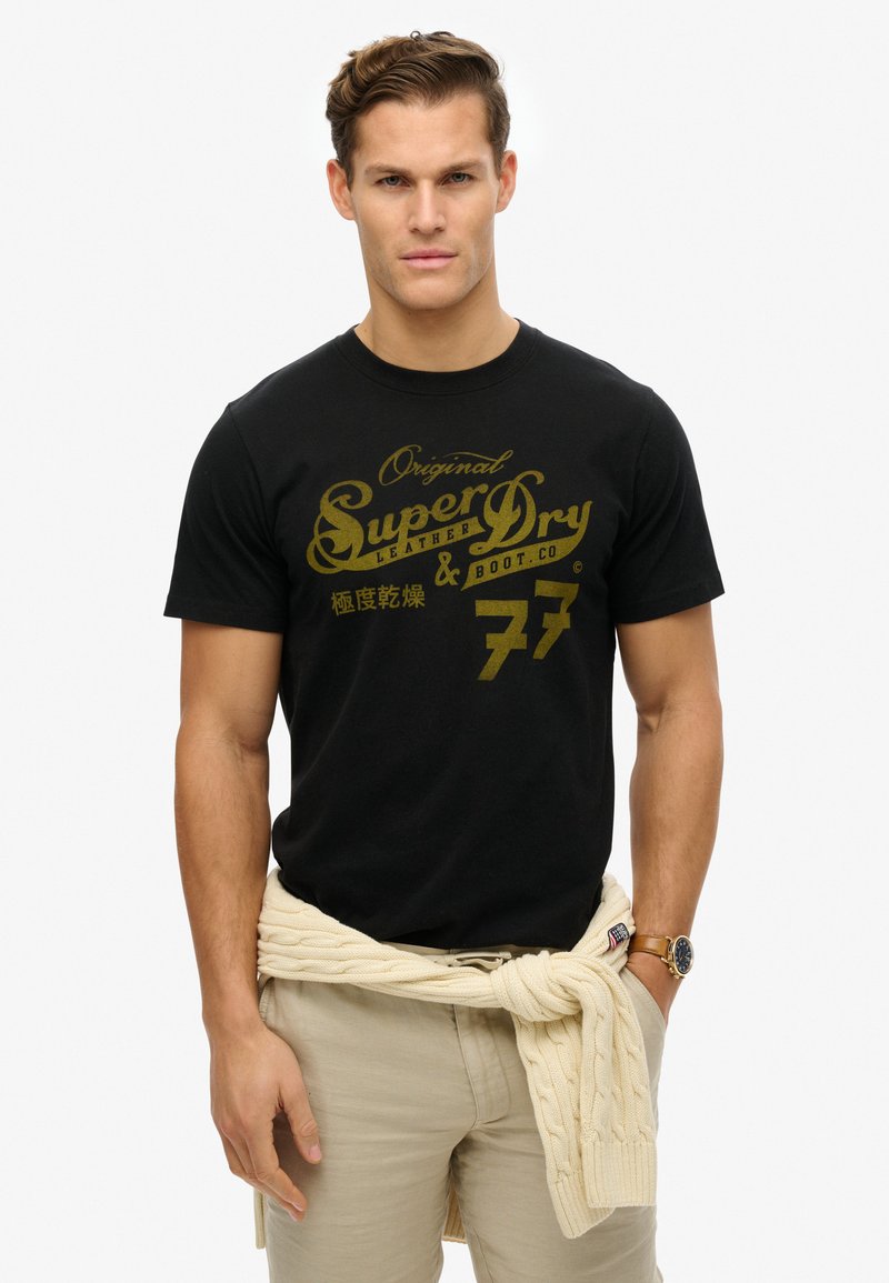 Sort bomulds T-shirt med guld grafisk tekst. Bæres med lysebeige bukser og en cremefarvet kabelstrikket sweater bundet om taljen.