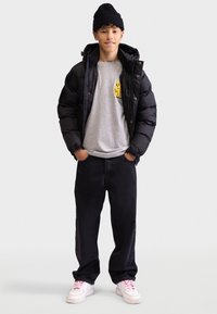 Schwarze Pufferjacke, graues T-Shirt mit gelbem Grafikdruck, lockere schwarze Jeans und weiße Sneaker mit rosa Schnürsenkeln. Eine gestrickte schwarze Mütze wird getragen.