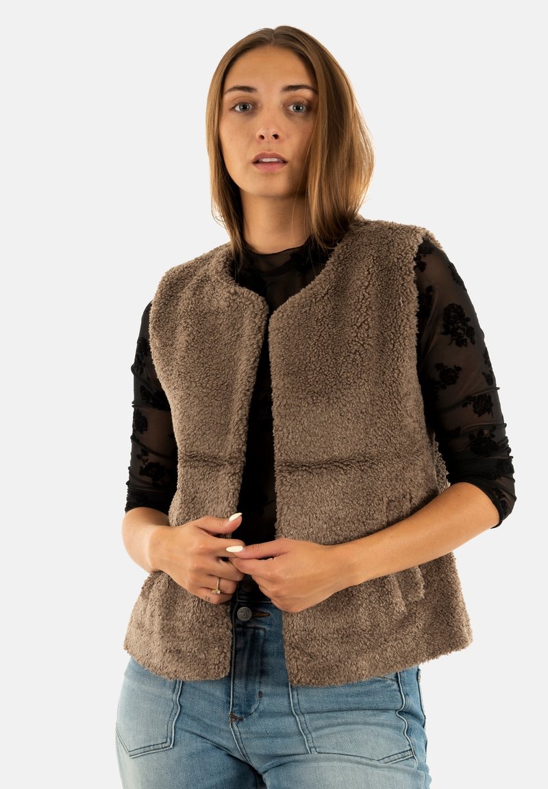 Giovane donna che indossa un gilet senza maniche marrone con una texture sopra una blusa nera trasparente a fiori e jeans azzurri chiari, mentre aggiusta il gilet con entrambe le mani.