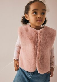 Ein junges Kind mit lockigem Haar trägt eine weiche rosa ärmellose Fleece-Weste, ein blumiges Langarmshirt und blaue Jeans und steht drinnen.